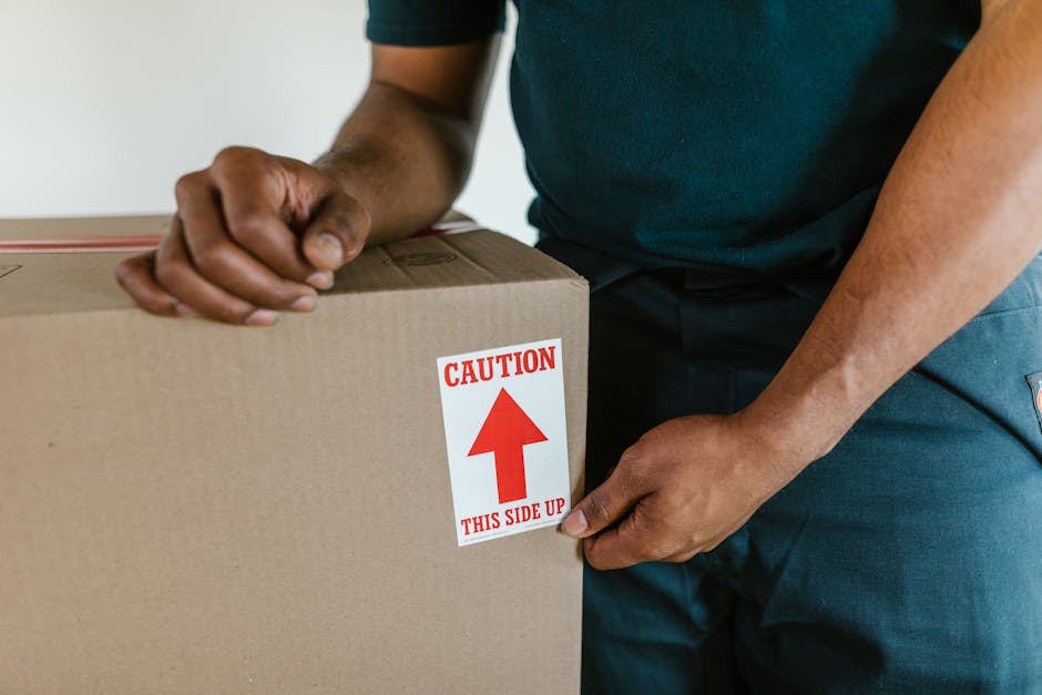 A person wearing a dark T-shirt is shown packing a fragile glass item into a white cardboard box lined with white packing peanuts to cushion the contents during home relocation or removals. The indivi
